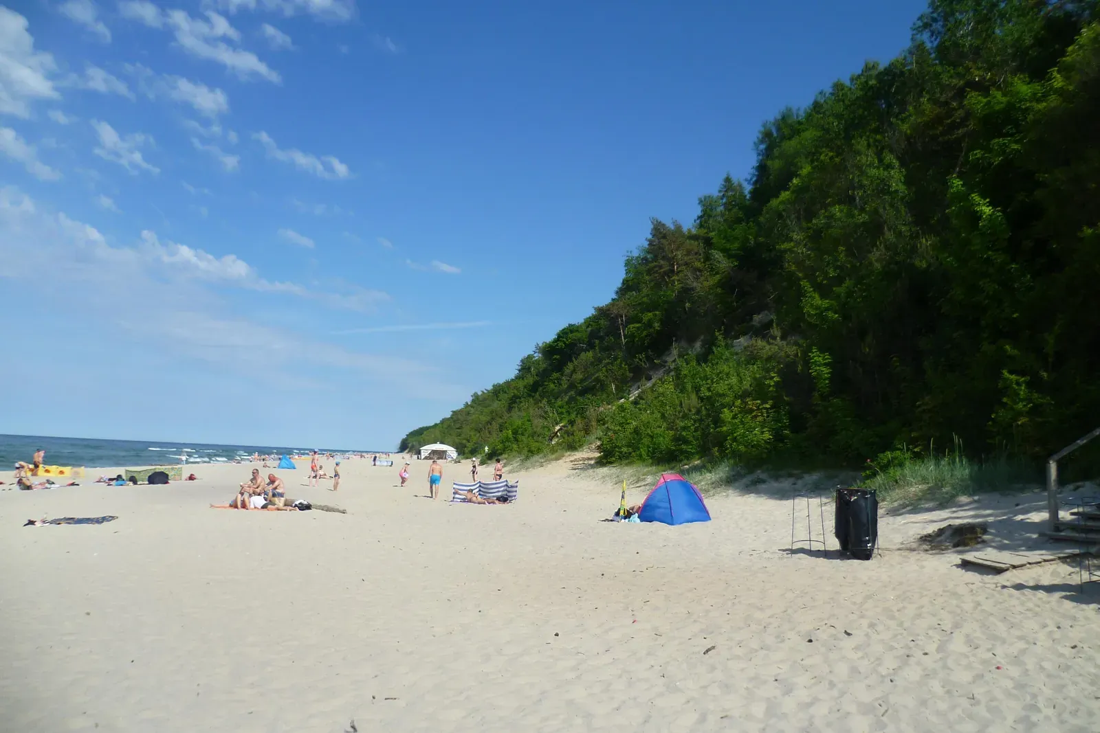 Szeroka plaża w Jastrzębiej Górze u stóp wysokiego, zalesionego klifu, w oddali plażowicze i morze