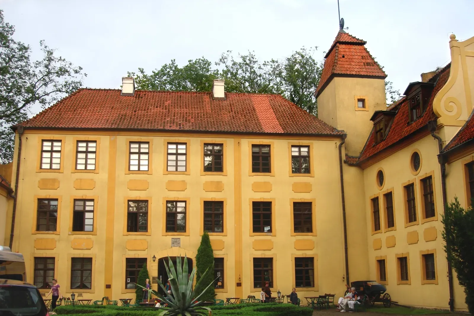 Żółta fasada zamku w Krokowej z czerwonym dachem i wieżą, przed wejściem dziedziniec z ogrodem i agawą